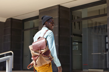 Cheerful handsome dark-skinned male tourist with backpack wearing trendy clothing about to enter modern building of embassy to extend visa while spending summer vacations in foreign country