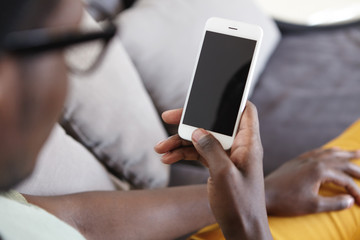 Cropped indoor image of unrecognizable dark-skinned man relaxing on couch in living room, using home wi-fi on modern mobile phone with blank copy space screen for your text or advertising content