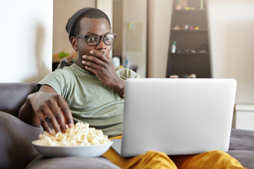 Fototapeta na wymiar Funny young dark-skinned man sitting on grey sofa in living room with notebook pc on his lap, staring at screen with shocked or scared look, covering mouth with hand while watching scary movie