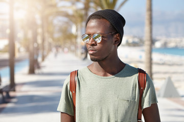 Confident serious African American 20-year old man with backpack walking along street on summer sunny day. Black hipster in hat and mirroerd lens sunglasses going to beach at daytime in resort town