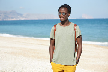 Handsome happy young black male tourist with backpack dressed in stylish clothing standing on pebble beach with blue sea and sky in horizon, waiting for friends to have nice walk along shore