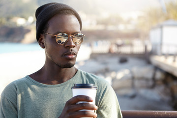Outdoor summer shot of handsome fashionable African American male hipster feeling relaxed and carefree, drinking coffee out of paper cup, waiting for girlfriend on city street bench in urban setting