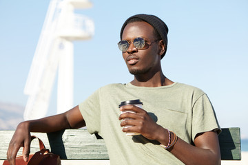 Summer, holiday and vacation. Outdoor shot of relaxed carefree dark-skinned male tourist in eyewear and hat sitting on wooden bench, holding paper cup with hot drink, lifeguard stand in background