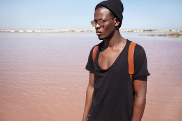 People, lifestyle, travel and fashion concept. Attractive fashionable young African American male model wearing black low-neck t-shirt, hat and mirrored lens shades posing outdoors by the sea
