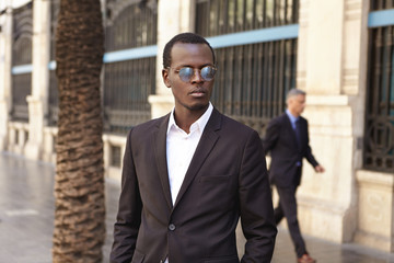 Outdoor portrait of handsome serious young black European businessman wearing round sunglasses and black suit waiting for taxi outside modern office building while hurrying to business meeting