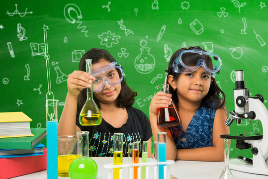 2 Cute Little Indian Girls Doing Science Experiment Or Project In A Classroom With Green Chalkboard Having Science Doodles