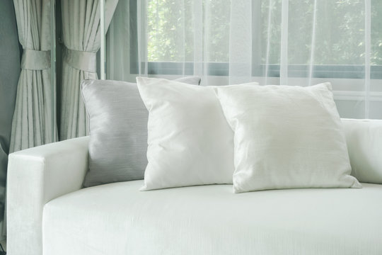 Pillows lay on white sofa with sheer curtain in background