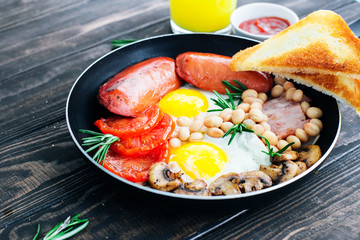 Traditional English breakfast eggs, tomatoes, bacon, sausages, mushrooms, toast and beans decorated with rosemary and orange juice on a wooden background