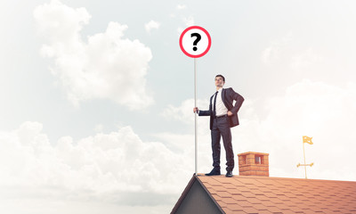 Businessman on brick house roof showing banner with question mar