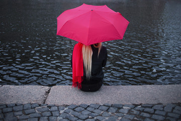 Girl in a bright pink scarf, rubber boots and umbrella sitting on the banks of the river, back. Grey grim landscape.