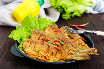 Fried fish carp (sazan) on a cast-iron frying pan with lettuce leaves on a dark wooden background.