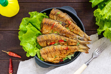 Fried fish carp (sazan) on a cast-iron frying pan with lettuce leaves on a dark wooden background. The top view.