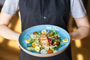 Vegetable salad with cucumber, lettuce, cherry tomatoes, salmon and eggs. Waitress holding dish in hands