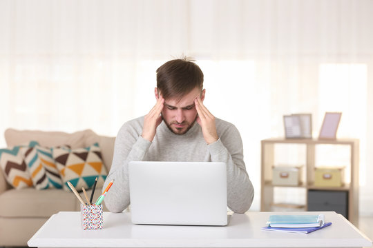 Young man with headache sitting at table with laptop