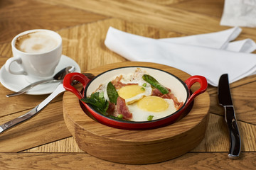 bacon and eggs with green peas in red pan and cup of americano coffee on wooden table in restaurant