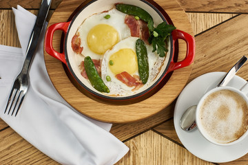 bacon and eggs with green peas in red pan and cup of americano coffee on wooden table in restaurant
