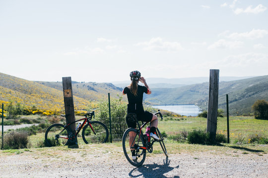 female cyclist in all black making a photo of beautiful view in front of a wood pillar fence and red bicycle sitting in the background, lake and green rolling hills.
