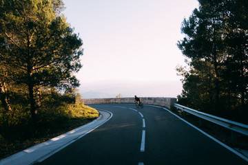 Tiny female cyclist dressed in black descending narrow road in spainish mountains haze off in the distance surrounded by tall green trees during pink, yellow, white light sunset.