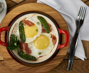 bacon and eggs with green peas in red pan served on wooden table in restaurant