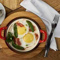 bacon and eggs with green peas in red pan served on wooden table in restaurant