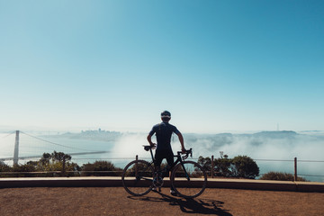 Fototapeta premium Young fit skinny male cyclist dressed in dark clothing sits on bike, bicycle looking over blueish foggy San Francisco, california in the dirt with gaurd rails and empty sky.