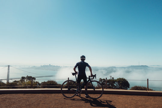 Young fit skinny male cyclist dressed in dark clothing sits on bike, bicycle looking over blueish foggy San Francisco, california in the dirt with gaurd rails and empty sky.