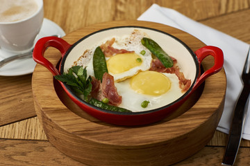 bacon and eggs with green peas in red pan and cup of americano coffee on wooden table in restaurant