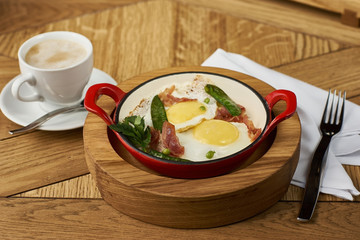 bacon and eggs with green peas in red pan served on wooden table in restaurant