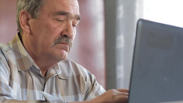 An elderly man with a mustache sits behind a laptop and solves problems. He looks seriously at the monitor