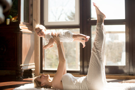 Young yogi smiling mother doing yoga exercise at home, wearing white sportswear, holding high funny daughter, enjoying activities with baby, fun and sport practice. Healthy lifestyle concept photo
