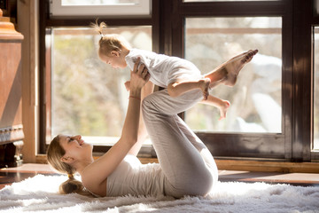 Young mother working out wearing white sportswear,  exercising at home with baby daughter, leg...