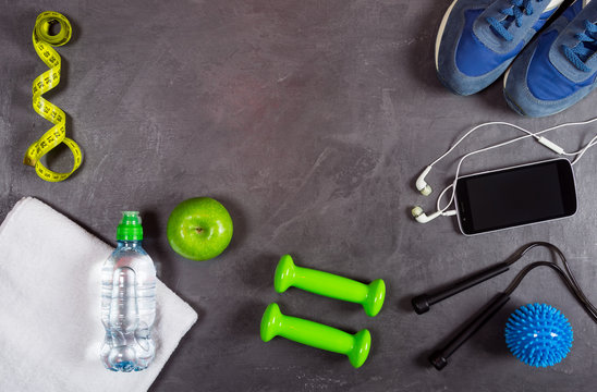 Fitness equipment background. Dumbbell, apple, water bottle, towel, smartphone and headphones, measuring tape, sneakers on grey background. Top view