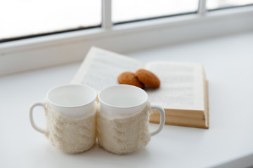 Two white mugs in a scarf stand on a table in the background of a window.
