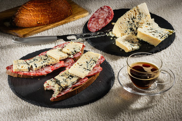 Two sandwiches from black bread with cheese of Dorblu and raw smoked sausage on black slate plates, a cheese knife, a wooden chopping board with bread and a cup of hot coffee on a transparent saucer