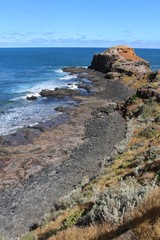 Walk to the Pulpit Rock along black lava stones and red soil contrasting with the blue ocean and sky at Cape Schank on the Mornington Peninsula, Australia