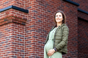 Fototapeta na wymiar Modern pregnant woman in a light jacket and casual dress posing against a brick house background