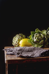 Baby ripping organic Artichokes in the rustic wooden board with lemon, solfetka. Dark background.