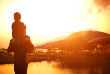 The boy sits on the shoulders of his father. Golden hour scene .