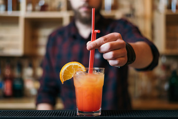 Handsome bearded barman is making tequila sunrise cocktail in night club. Selective focus.