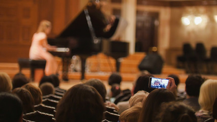 Spectators in concert hall during performing piano girl- people shooting performance on smartphone - obrazy, fototapety, plakaty