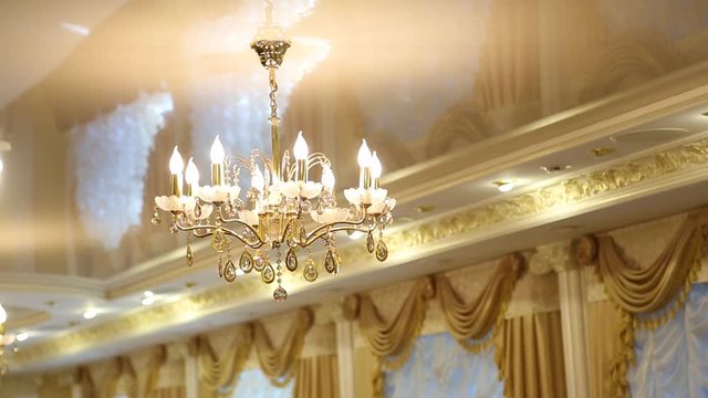 Luxury bright interior. Closeup of chandelier hanging from stretch golden color ceiling in living room. Windows decorated with beautiful draped curtains. Camera moves from left to right.
