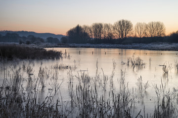 Beautiful vibrant English countryside lake image with frost and frozen lake in Winter at sunrise