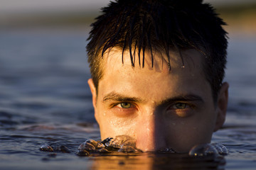 Portrait of young man sitting in the water at the sea. Half of face is covered with water, accent...