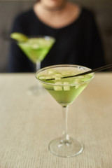 Two glasses of apple martini and apple slices on table in cafe. Young woman blurred at background