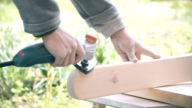 Close-up of hands grinding a wooden plank with an orbital detail sander outdoors on a sunny day. The plank material is pine wood.