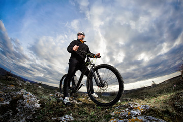 Plakat Photo below of the mountain cyclist in the black sportwear on the rocks against dramatic sky with clouds.