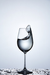 Pure splashing water wave in the wineglass while standing on the glass with drapes against light background.