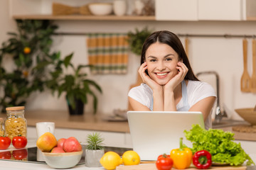 Obraz na płótnie Canvas Young Woman in kitchen with laptop looking recipes, smiling. Food blogger concept