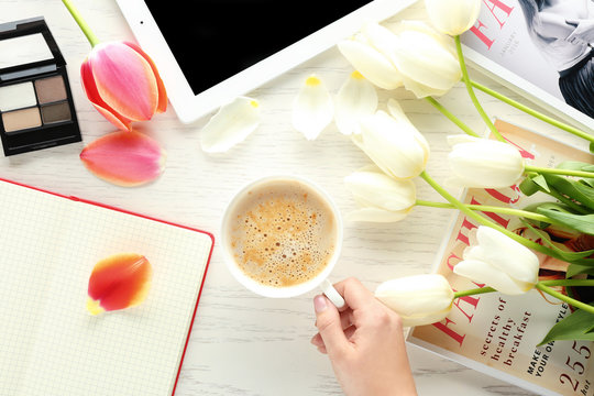 Female hand holding cup of coffee on table with flowers, notebook, magazines and cosmetics