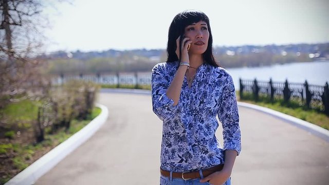 Beautiful Asian girl in jeans walking in summer in the park on a sunny day and talking on the phone